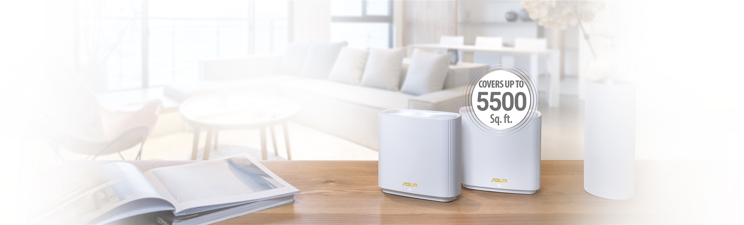 L'immagine mostra un ambiente domestico moderno e luminoso. In primo piano su un tavolo di legno ci sono due dispositivi ASUS, probabilmente router o dispositivi di rete, con un adesivo che indica che coprono fino a 5500 piedi quadrati. Sullo sfondo si vede un soggiorno con un divano, un tavolino e una grande finestra. C'è anche un libro aperto sul tavolo vicino ai dispositivi. L'intera scena appare accogliente e ben illuminata.